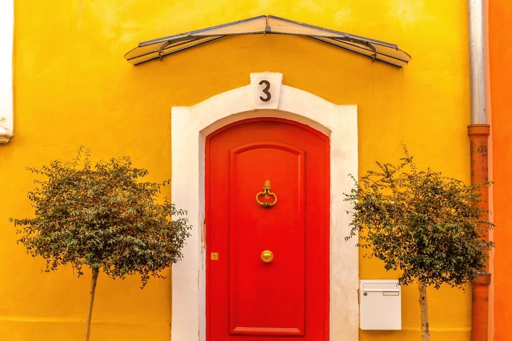 A red front door numbered 3 in a sunny yellow wall, with two small trees